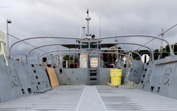 The image shows the open bow ramp of a military landing craft vessel, viewed from the ramp looking toward the stern. The vessel is painted gray and features a wide, ribbed metal ramp designed for vehicle and troop offloading. The craft appears to be docked, with trees and overcast skies visible in the background.