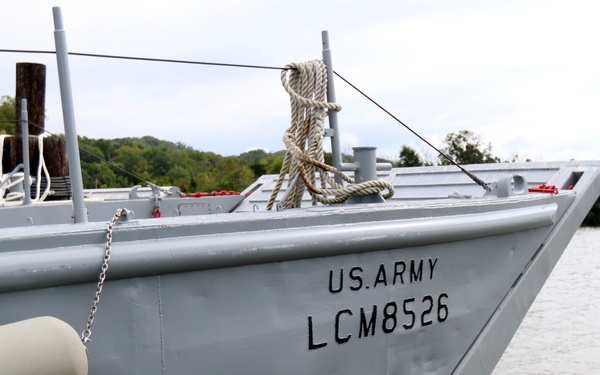 The image shows a U.S. Army Landing Craft Mechanized (LCM) vessel designated LCM8526, moored on a calm waterway. The gray military vessel is viewed from a close angle showing its bow and port side, with wooded shoreline visible in the background. The craft appears to be docked or anchored near a pier in overcast daylight conditions.