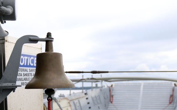 The image shows a brass ship's bell mounted on a gray metal bracket on the exterior of a vessel, with a red braided rope clapper cord hanging below it. The bell is positioned near the top of what appears to be a boarding ramp or gangway leading down to a lower deck area. A partially visible 'NOTICE' sign referencing Material Safety Data Sheets is mounted on the same structure.