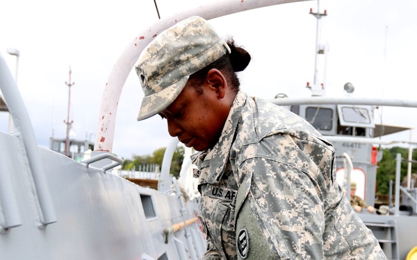 A U.S. Army soldier in ACU (Army Combat Uniform) digital camouflage is performing maintenance or inspection work on a military vessel, using a wrench or similar hand tool on a fitting mounted to the ship's hull. The soldier is wearing a patrol cap and has a name tape reading 'U.S. AR[MY]' visible on the uniform. The setting appears to be a military watercraft facility or dock.