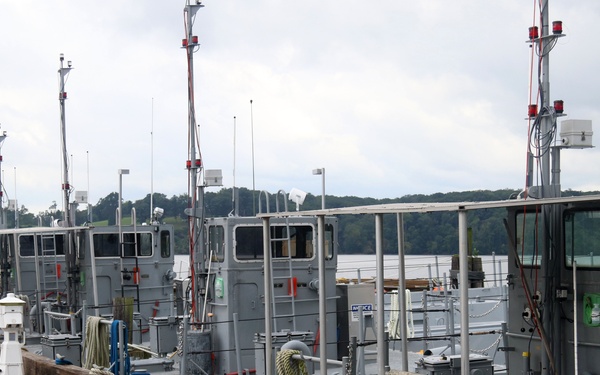 A wooden dock or pier runs alongside several gray military or government patrol vessels moored in a calm waterway. The boats are uniformly painted gray and equipped with antennas, navigation lights, cameras, and various deck equipment. Wooded shoreline and an overcast sky are visible in the background.