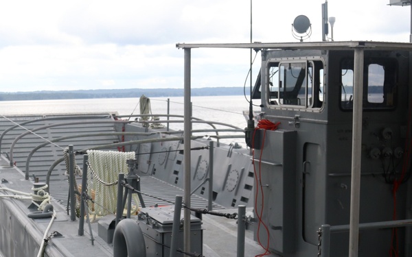 A gray military or government utility vessel is docked at a wooden pier on a calm body of water. The flat-decked workboat features a pilothouse, various deck equipment, mooring lines, and operational gear. The overcast sky and distant tree-lined shoreline suggest an inland waterway or estuary setting.
