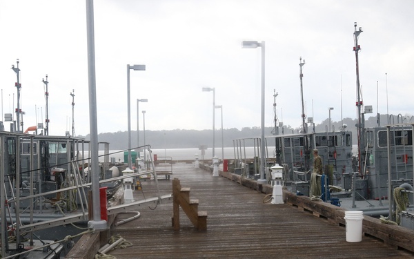 A wet wooden dock or pier stretches into the distance under overcast, misty conditions. Multiple gray military or utility vessels are moored on both sides of the dock. The scene appears to be at a military or government marina, with the wet planking suggesting recent rainfall.
