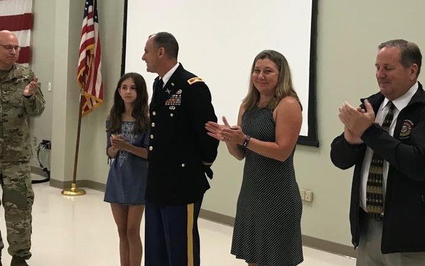 A military ceremony appears to be taking place in an indoor facility, likely a promotion or retirement event. A U.S. Army officer in dress uniform stands at center with what appears to be family members and colleagues applauding. An American flag is visible in the background alongside a projection screen.