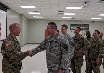 A group of U.S. Army soldiers are gathered in what appears to be a military briefing room or assembly hall. Two soldiers in the foreground are shaking hands and smiling, suggesting a formal recognition or greeting ceremony. Several other soldiers stand in the background observing the interaction.