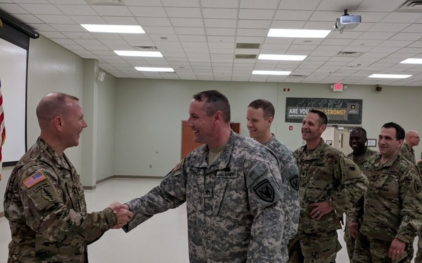 A group of U.S. Army soldiers are gathered in what appears to be a military briefing room or assembly hall. Two soldiers in the foreground are shaking hands and smiling, suggesting a formal recognition or greeting ceremony. Several other soldiers stand in the background observing the interaction.