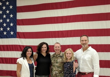 A group of five people pose for a photo in front of a large American flag displayed on an indoor wall. One individual is wearing U.S. Army camouflage (OCP/MultiCam) uniform, suggesting a military event such as a deployment ceremony, homecoming, or promotion. The group appears to be family or close friends gathered to celebrate or support the service member.