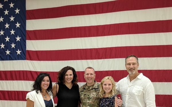 A group of five people pose for a photo in front of a large American flag displayed on an indoor wall. One individual is wearing U.S. Army camouflage (OCP/MultiCam) uniform, suggesting a military event such as a deployment ceremony, homecoming, or promotion. The group appears to be family or close friends gathered to celebrate or support the service member.