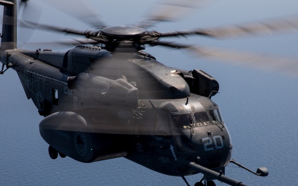 A large dark gray military helicopter marked with the number '20' is photographed in flight over open ocean. The helicopter has an aerial refueling probe extended from its nose, with a drogue basket (parachute-stabilized refueling receptacle) visible at the end of a hose. The image appears to capture an in-flight refueling operation, likely involving a CH-53E Super Stallion or similar heavy-lift helicopter.