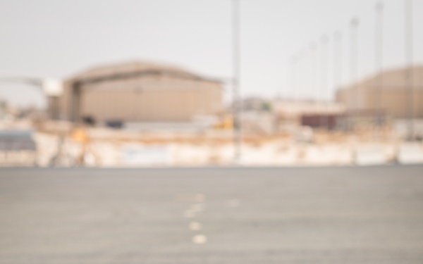 A military personnel member in a tan/desert-colored flight suit sits on what appears to be the open ramp or cargo door of a military aircraft, facing away from the camera toward a tarmac or airfield. The individual is wearing green aviation-style communication headset with a boom microphone. The background shows arid, desert-environment airfield infrastructure including hangars and utility poles.