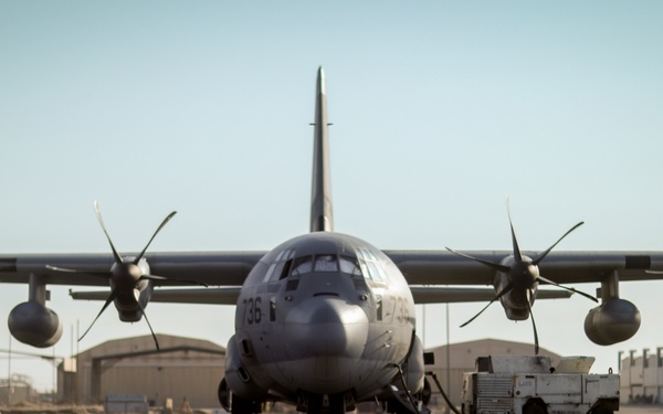 A military turboprop transport aircraft, identifiable as a C-130 Hercules variant, is parked on a flight line at what appears to be a desert air base. The aircraft bears the tail number '036' and is surrounded by ground support equipment and personnel. The arid environment and tan-colored hangars in the background suggest a Middle Eastern or similarly dry-climate military installation.