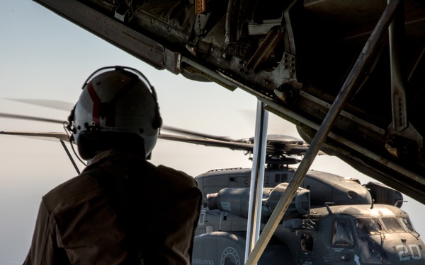 A military aircrew member wearing a flight helmet and communications headset sits at the open rear ramp of a large military aircraft, observing a CH-53E Super Stallion heavy-lift helicopter flying in close formation. The helicopter, marked with the number '20', is visible in the background against a hazy sky. The scene appears to depict an aerial operation, possibly a formation flight or air-to-air refueling observation.