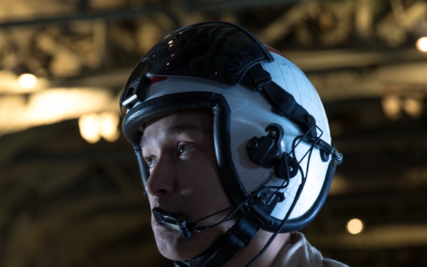 A military aviator or flight crew member is photographed wearing a white flight helmet with integrated communications equipment and a boom microphone. The individual is dressed in a flight suit and appears to be inside what looks like a naval vessel or aircraft hangar, illuminated by warm ambient lighting. The portrait-style image captures the subject in a three-quarter profile view with a focused, alert expression.