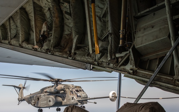A military personnel member is lying prone on the open ramp of a large military aircraft, observing a Navy helicopter conducting an aerial refueling operation. The helicopter, marked 'NAVY' with the number '20', is connected to a refueling drogue extended from the aircraft. The scene takes place at altitude over what appears to be open water or hazy sky.