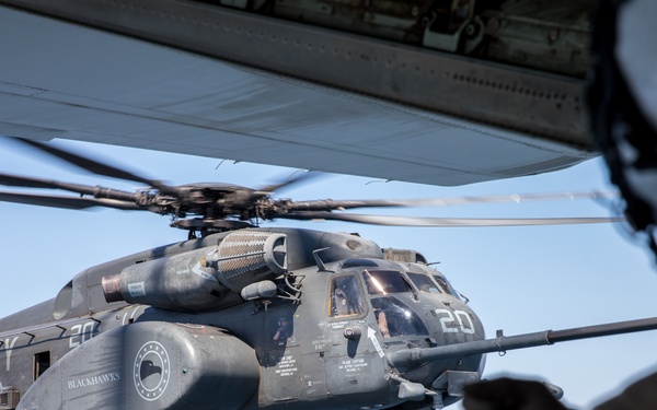 A U.S. Navy MH-53E Sea Dragon helicopter, marked with the number '20' and squadron designation 'HM-15 Blackhawks,' is hovering in flight. A crew member in the foreground, wearing a flight helmet and gloves, is pointing toward the helicopter, likely during a coordination or signaling operation. The scene appears to be an aerial or shipboard operation conducted in clear daylight conditions.