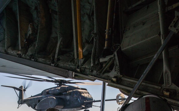 A military crew member lies prone on the open rear ramp of a large transport aircraft, wearing a flight helmet with a red cross/stripe marking, observing or managing an aerial refueling operation. A large military helicopter, consistent with a Sikorsky CH-53 Sea Stallion or similar heavy-lift helicopter, is visible flying in close proximity off the ramp. A refueling drogue basket is visible between the two aircraft, indicating an active air-to-air refueling evolution.