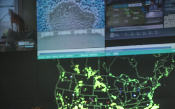 Three military personnel in olive green flight suits are working in what appears to be an operations or command center. A large display wall shows a glowing green map of the continental United States with what appears to be air traffic or network data overlaid. Multiple computer workstations are visible throughout the room.