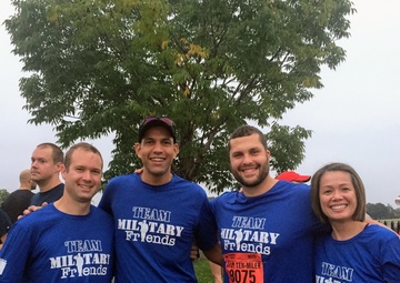 Four runners wearing matching blue 'Team Military Friends' t-shirts pose together at what appears to be the Army Ten-Miler race event. They are displaying race bibs with numbers visible including 2391, 2852, 8075, and 22494. The group appears to be in good spirits, smiling and giving thumbs up, likely before or after the race.