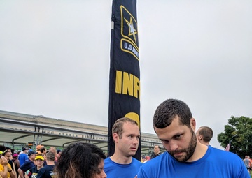 A group of participants wearing blue 'Team Military Friends' shirts are gathered at what appears to be an Army-sponsored running event, likely the Army Ten-Miler based on the bib number and date visible. A woman is writing on another participant's arm with a marker, a common race-day practice for noting bib numbers or pace goals. The event takes place outdoors near a large U.S. Army Infantry banner, with many other runners visible in the background.