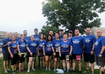 A large group of approximately 20 people are posing together outdoors wearing matching blue 'Team Military Friends' t-shirts. Several participants are wearing race bibs with visible numbers, suggesting this is a running event, likely the Army Ten-Miler based on the bib designs. The group is standing on a grassy area with a large building and trees visible in the background.