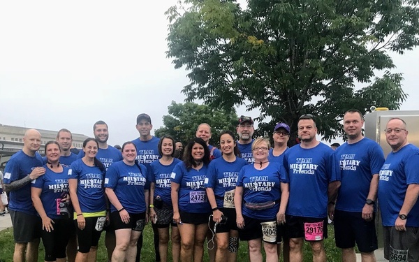 A large group of approximately 20 people are posing together outdoors wearing matching blue 'Team Military Friends' t-shirts. Several participants are wearing race bibs with visible numbers, suggesting this is a running event, likely the Army Ten-Miler based on the bib designs. The group is standing on a grassy area with a large building and trees visible in the background.
