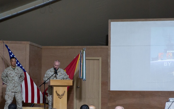 A military ceremony is taking place in an auditorium or assembly hall, with a speaker at a podium addressing a large audience of uniformed personnel. A projection screen displays text reading '243rd Navy Birthday - Forged by the Sea,' indicating this is a U.S. Navy birthday celebration. Two military personnel stand on stage near flags including the American flag and what appears to be a Marine Corps flag.