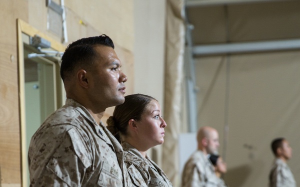 Four military personnel in desert camouflage uniforms (MARPAT pattern consistent with U.S. Marines) stand at attention during what appears to be a formal ceremony or celebration. A table in the foreground is set with white plates, decorated cakes, and patriotic decorations in red, white, and blue. The setting appears to be an indoor military facility or forward operating base.