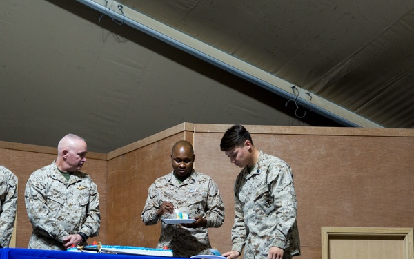 Four military personnel in desert camouflage uniforms (MARPAT pattern, consistent with U.S. Marines) are gathered on a stage around a table covered with a blue tablecloth. A cake is visible on the table, suggesting this is a military birthday or commemorative ceremony. A dark blue flag with gold fringe, consistent with a U.S. Navy or Marine Corps unit flag, is displayed on the right side of the stage.