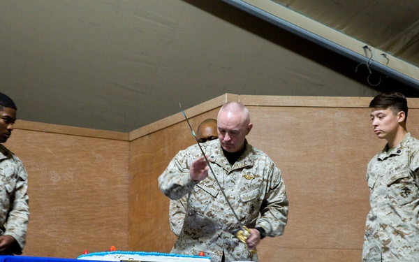Three military personnel in desert camouflage uniforms are gathered around a table with a blue tablecloth during what appears to be a military cake-cutting ceremony. The central figure, a senior officer, is using a ceremonial sword to cut a white cake on the table. This is consistent with a U.S. Marine Corps or Navy birthday or commemorative celebration tradition.