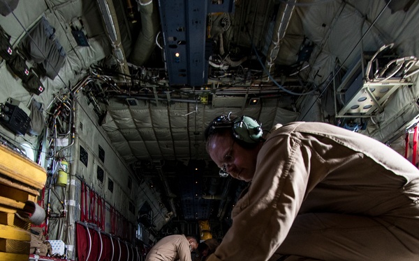 Two military personnel in tan/khaki uniforms are working inside the cargo hold of a large military transport aircraft, likely a C-130 Hercules or similar aircraft. The foreground individual is crouching and manipulating floor-mounted cargo rail hardware, while a second person is visible further back in the fuselage in a prone or kneeling position. The interior shows typical military cargo aircraft features including tie-down rails, structural ribs, wiring, and red cargo restraint systems.