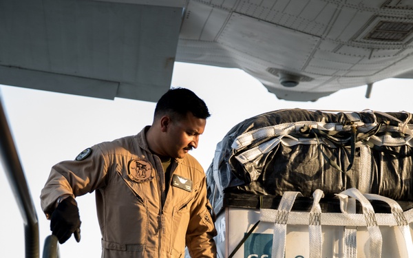 A military aircrew member in a tan flight suit is loading or securing a cargo pallet aboard what appears to be a military transport aircraft. The pallet is secured with white cargo netting and straps, and bears a label reading 'GS Contract Holder.' The scene appears to be taking place on a flight line during daylight hours in a warm, arid environment.