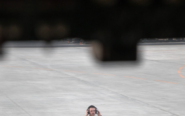 The image is taken from inside a military aircraft cockpit, looking through the windscreen at a ground crew member standing on a flight line or tarmac. The crew member is wearing a tan/khaki flight suit and holding communication headphones to their ears, connected by a cord to the aircraft. Green cockpit instrument displays are visible in the foreground, blurred due to depth of field.