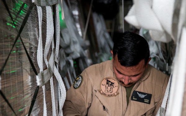 A U.S. Marine Corps Staff Sergeant named Lopez is working inside what appears to be a military cargo aircraft, securing or adjusting cargo straps and tie-downs. He is wearing a tan flight suit with a squadron patch and a name tag identifying him as 'SSGT Lopez, USMC' with aviator wings. The cargo hold is densely packed with palletized cargo secured with white netting and straps.