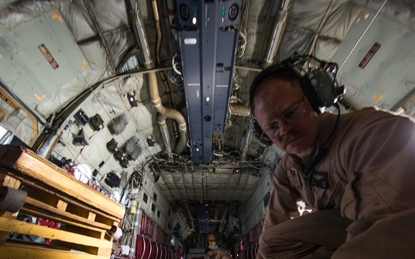 A military aircrew member in tan/desert flight suit is working inside the cargo hold of a large military transport aircraft, likely a C-130 Hercules. The individual is wearing aviation headset and glasses, and appears to be inspecting or adjusting cargo floor rails. The wide-angle fisheye lens captures the full length of the aircraft's interior, revealing wooden cargo pallets and standard military cargo loading equipment.