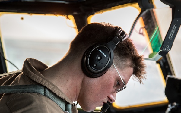 A young pilot or aircrew member is seated in what appears to be a military or large transport aircraft cockpit, reviewing documents or a checklist. They are wearing a tan/khaki flight suit and Bose noise-canceling headphones with an attached boom microphone. The cockpit displays modern avionics including digital instrument panels visible on the right side.