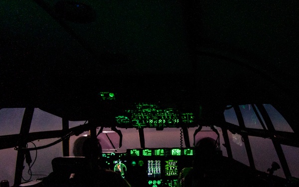 A military aircraft cockpit photographed at night, illuminated predominantly by green instrument lighting consistent with night vision goggle (NVG) compatible lighting. Two crew members are visible in silhouette seated at the flight deck, surrounded by extensive avionics panels. The wide cockpit layout and instrument configuration is consistent with a large military transport aircraft, possibly a C-130 or similar platform.