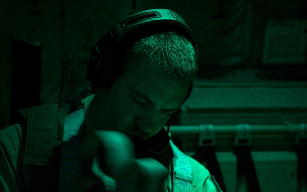 A military or aviation personnel member is seated inside what appears to be a military aircraft, illuminated by green lighting. The individual is wearing a flight suit, tactical vest, and communications headset, and appears to be handling or adjusting equipment. The green-tinted lighting suggests a night operations or red/green light environment typical of military transport aircraft.