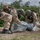 Two military personnel wearing CBRN (Chemical, Biological, Radiological, Nuclear) protective gear, including gas masks, helmets, desert camouflage MOPP suits, and rubber overboots, are conducting what appears to be a CBRN decontamination or casualty extraction drill. They are handling a clear plastic body bag or decontamination bag containing what appears to be a simulated casualty on a dirt surface. A red powder or paint mark is visible on the ground to the left, likely a training marker.