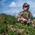 A male soldier in full tactical gear is positioned in a low crouch among grass and earthen terrain, holding an assault rifle. He is equipped with a combat helmet fitted with night-vision mount hardware, tactical communications headset, ballistic sunglasses, and a plate carrier vest. The outdoor setting appears to be a training or operational area with open sky and distant structures visible in the background.