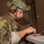 A male soldier in full tactical gear is focused on operating a small electronic device at a table. He is wearing a multicam camouflage uniform, tactical vest, combat helmet with mounted accessories, and tactical communications headset with boom microphone. The indoor setting appears to be a building interior with a brown wall and door visible in the background.