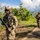 A group of heavily armed and equipped military personnel are conducting a patrol along a paved path in a tropical, vegetated environment. The lead soldier is facing the camera while holding an assault rifle, wearing full tactical gear including a helmet, plate carrier, and communications equipment. At least three additional soldiers are visible in the background moving along the same path.