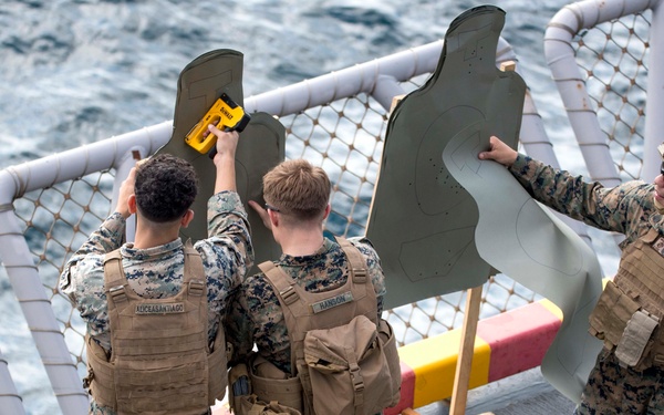 Three U.S. Marines in digital camouflage uniforms and tactical vests are handling human-silhouette paper shooting targets on what appears to be the deck of a naval vessel. They appear to be setting up or examining the targets, likely for a marksmanship qualification or training exercise at sea. The ocean is visible in the background beyond the ship's railing.