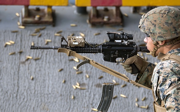 A U.S. Marine in full combat gear is actively firing a rifle, with numerous brass shell casings visibly ejecting and scattered across the deck surface. The shooter is positioned in a firing stance on what appears to be the deck of a naval vessel. The image captures a live-fire training exercise with exceptional detail of the ejected casings mid-air.