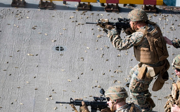 The image shows a group of military personnel, appearing to be U.S. Marines based on their distinctive MARPAT (digital camouflage) uniforms, conducting a tactical live-fire exercise on what appears to be the deck of a naval vessel. Multiple spent brass casings are scattered across the deck surface, indicating active or recent weapons discharge. The personnel are equipped with full combat gear including plate carriers, helmets, and M4-style carbines.