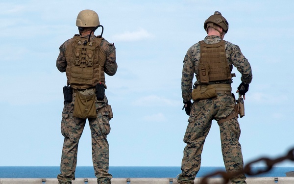 A group of military personnel in full combat gear stand at the edge of what appears to be a naval vessel's deck, facing the open ocean. They are wearing digital camouflage uniforms with tactical vests and backpacks. Heavy anchor chains are visible in the foreground, confirming a shipboard setting.
