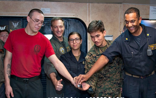 A group of military personnel, including Navy and Marine Corps members, are gathered around a large decorated cake for a ceremonial cutting. The cake features the U.S. Navy seal and an American flag design, suggesting a Navy birthday or commissioning anniversary celebration. The event appears to be taking place aboard a naval vessel based on the interior surroundings.
