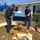 Two U.S. Coast Guard personnel are assisting with disaster cleanup efforts in front of a residential home. They are lifting a blue plastic storage container or cooler lid from a pile of debris that includes damaged carpeting and wood. The scene shows significant storm damage with fallen tree branches, a blue tarp on a nearby structure, and scattered debris across the yard.