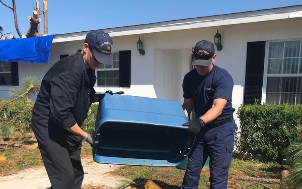 Two U.S. Coast Guard personnel are assisting with disaster cleanup efforts in front of a residential home. They are lifting a blue plastic storage container or cooler lid from a pile of debris that includes damaged carpeting and wood. The scene shows significant storm damage with fallen tree branches, a blue tarp on a nearby structure, and scattered debris across the yard.