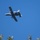 A military attack aircraft, identifiable as an A-10 Thunderbolt II (Warthog), is photographed in flight against a clear blue sky. The aircraft is captured in a banking maneuver, showing its underside with visible hardpoints and ordnance pylons. Tree foliage is visible in the lower portion of the frame, suggesting the photo was taken from ground level.