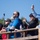 A group of spectators stand on a wooden viewing platform or bleachers, capturing photos and video of an off-screen event. Multiple people are holding up smartphones and a selfie stick with an action camera attached. The clear blue sky and casual attire suggest an outdoor daytime event.